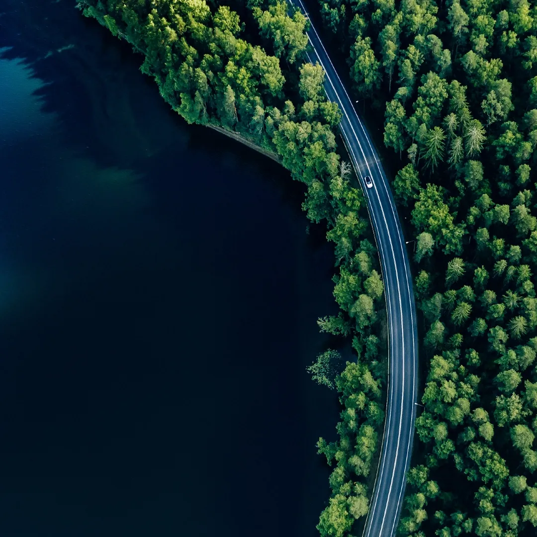 A car drives along a winding road between a forest and a lake.
