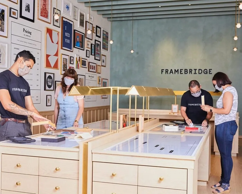 People wearing masks are working and discussing picture frames inside a stylish Framebridge store.
