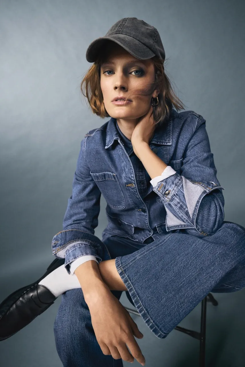 A woman in a denim jacket and jeans poses seated, wearing a dark cap and black shoes. (Image: Remy Farrell)