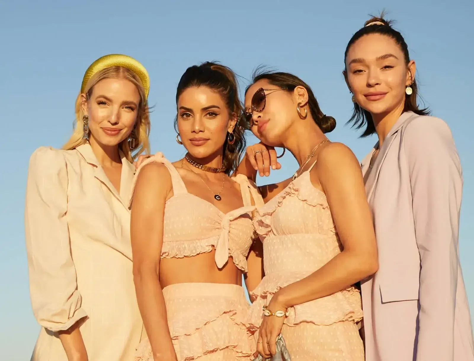 A group of four stylish women pose together in chic, summery outfits under a clear blue sky.