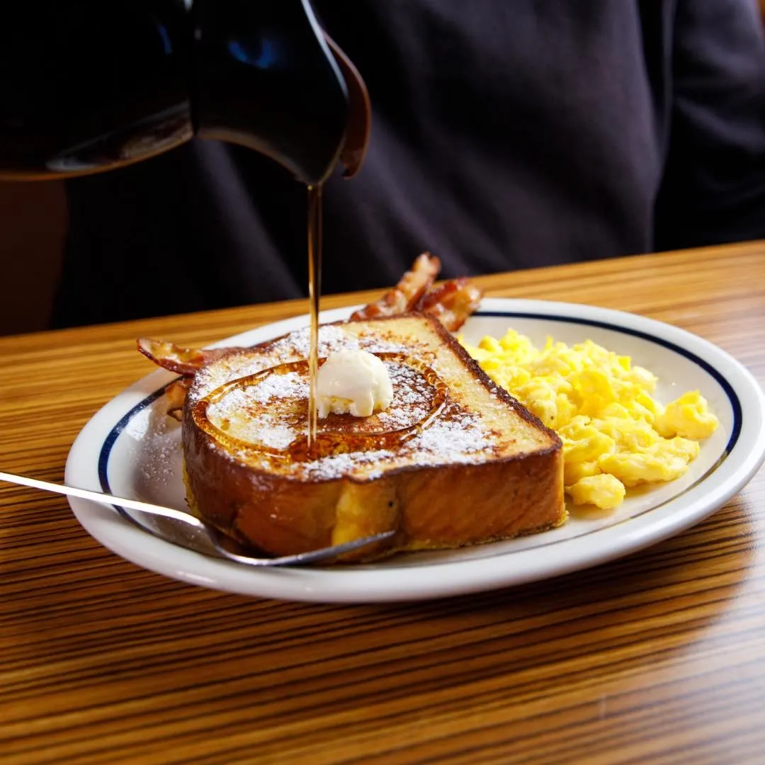 A plate of French toast with butter and syrup, served with scrambled eggs and bacon.