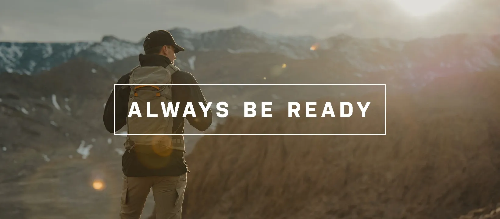A man wearing a backpack stands on a mountain overlooking a scenic landscape with the words 'ALWAYS BE READY' written across the image.
