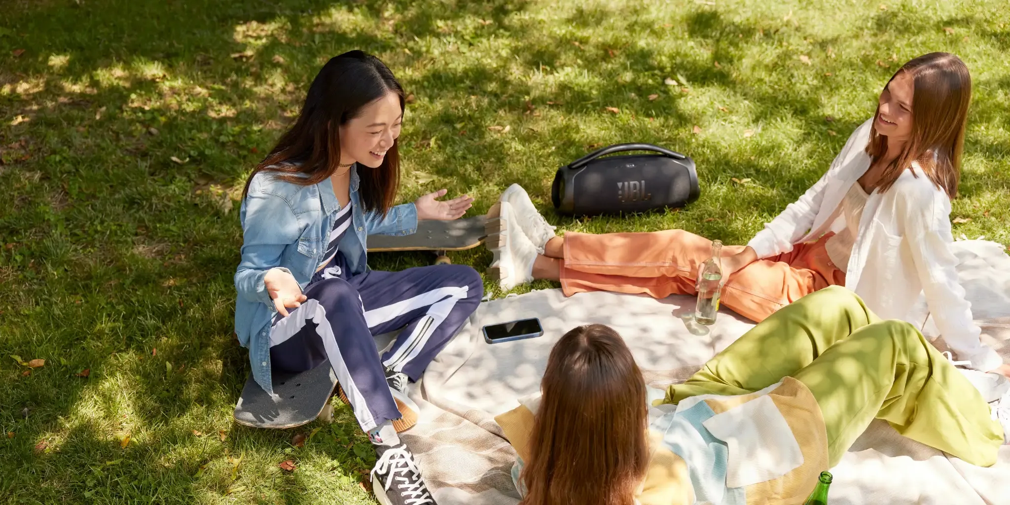 Three friends having a picnic with a JBL portable speaker in a park.