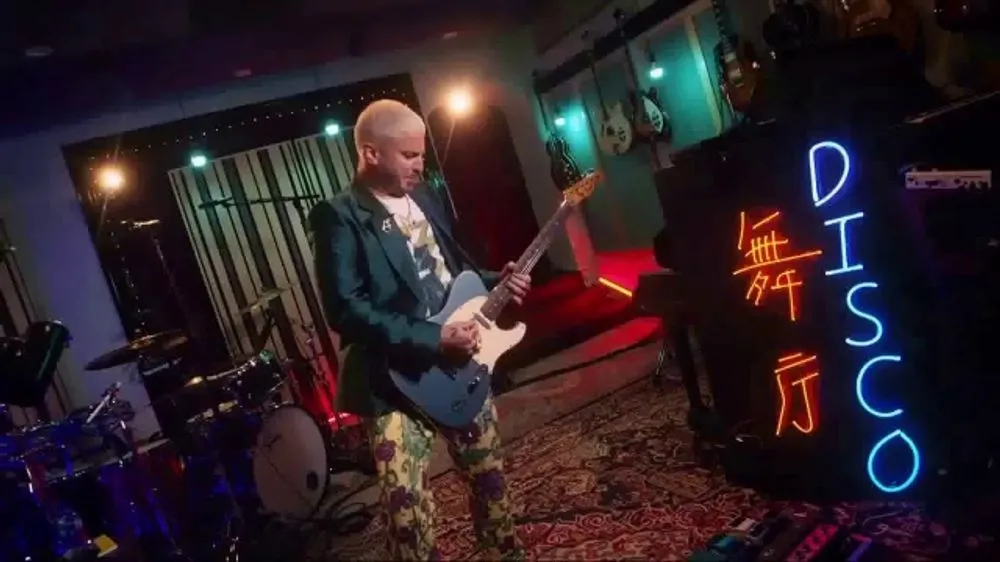 A musician plays an electric guitar in a colorful studio with neon lights and instruments around.