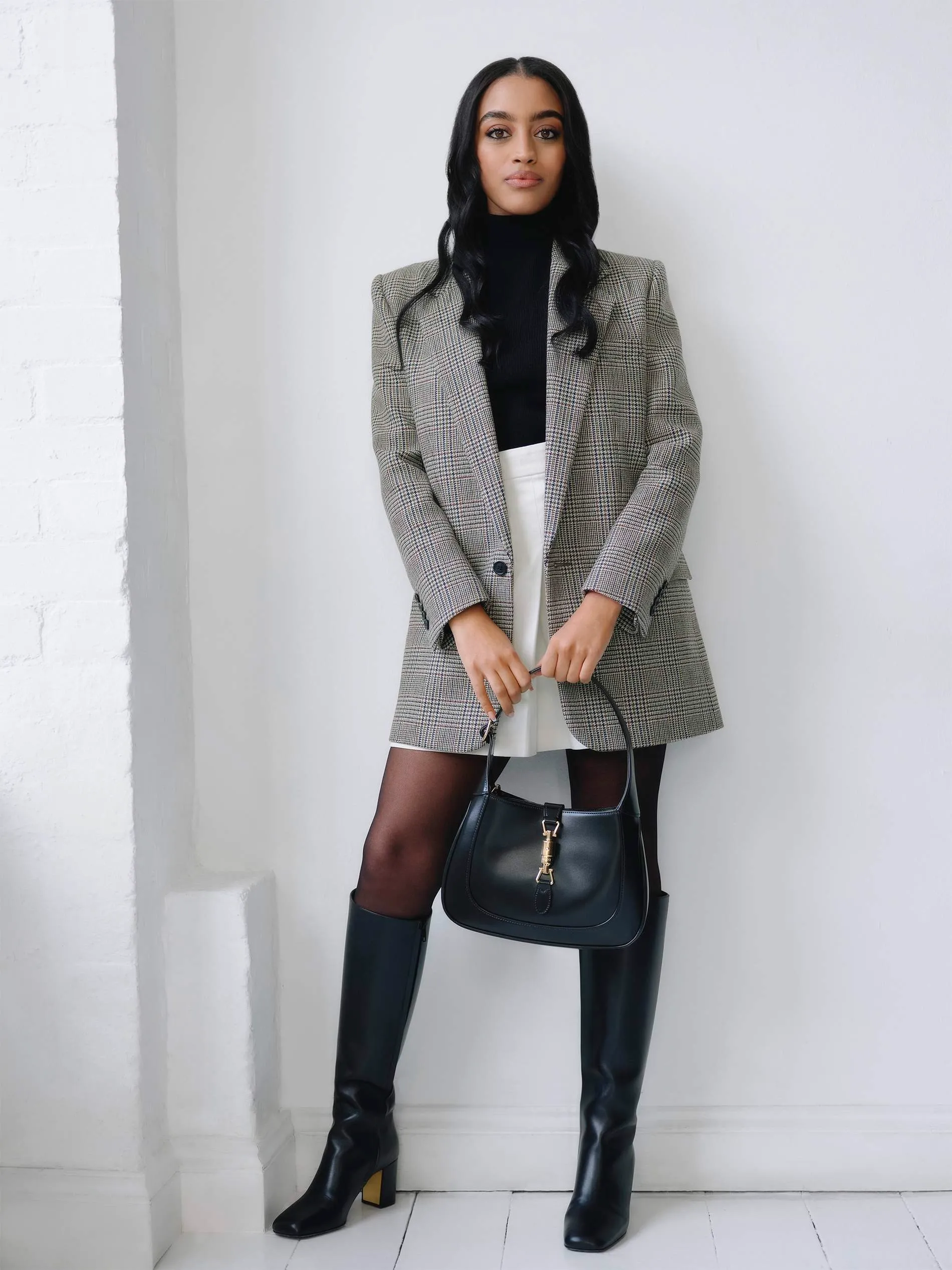 A stylish woman poses in a blazer, skirt, boots, and black handbag.