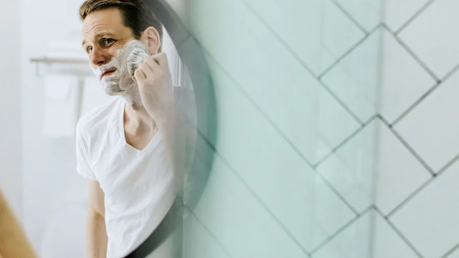 Man carefully shaving face in bathroom mirror with gentle technique for sensitive skin care